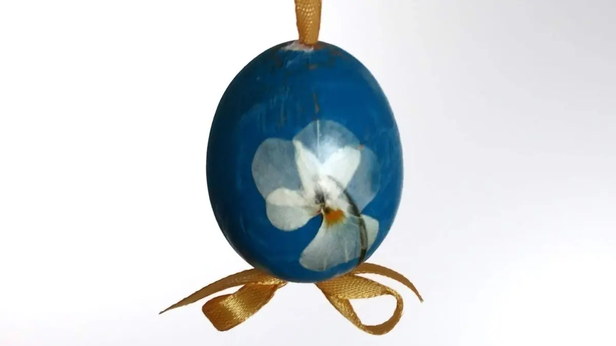 Ein blaues Osterei mit einer Bl&uuml;te h&auml;ngt dekorativ an einem gelben Band von einem Zweig, vor hellem Hintergrund.