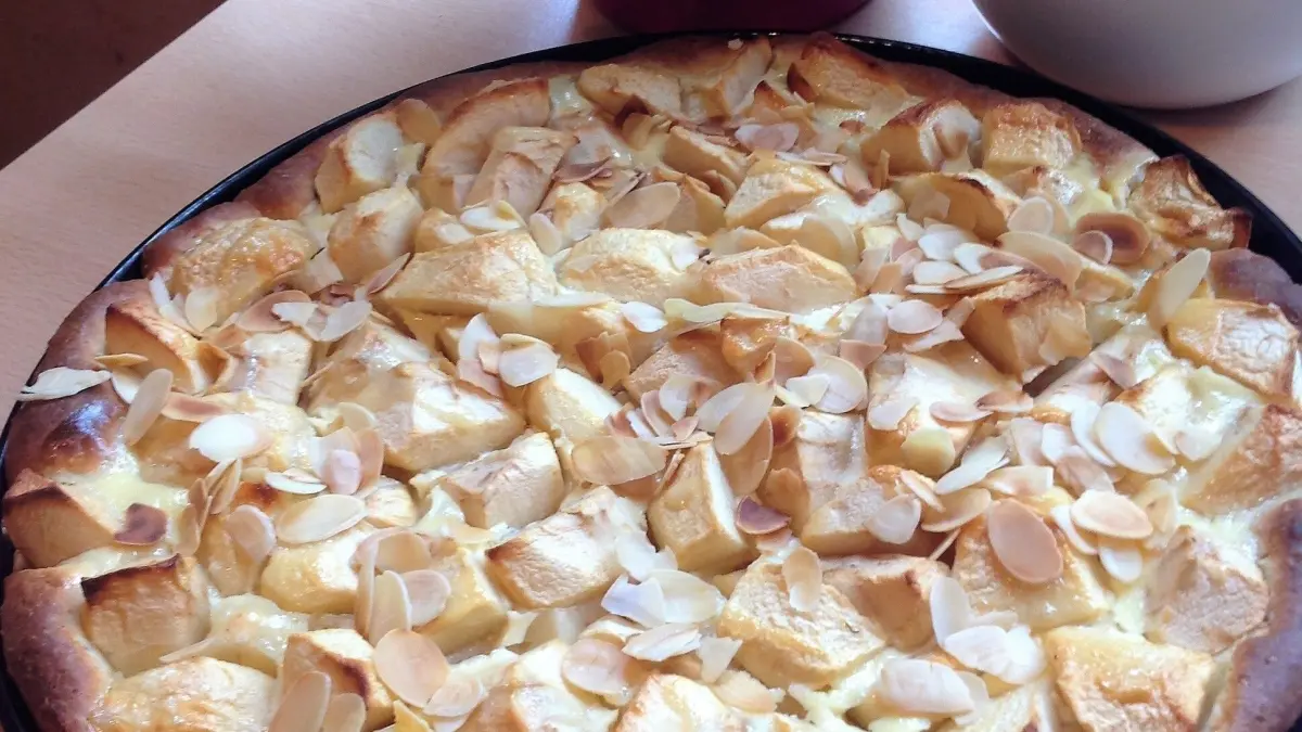 Der Hefe-Apfelkuchen mit Guss steht in einer runden Form und hat goldbraune Apfelst&uuml;ckchen auf der Oberfl&auml;che, umgeben von Pflanzen.