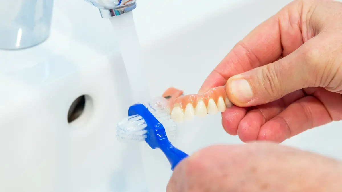 Zahnprothese wird mit einer Zahnb&uuml;rste gereinigt, w&auml;hrend sie &uuml;ber einem Waschbecken gehalten wird.