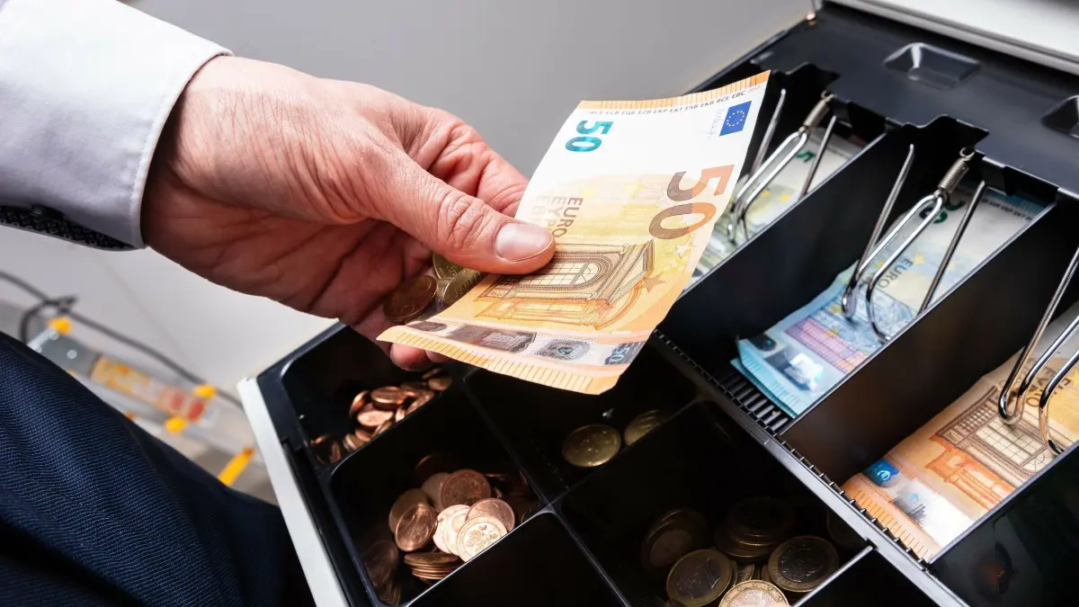 Eine Hand h&auml;lt einen 50-Euro-Schein, w&auml;hrend sie Geld abhebt, um eine Kasse in einem Einkaufsumfeld zu f&uuml;llen.