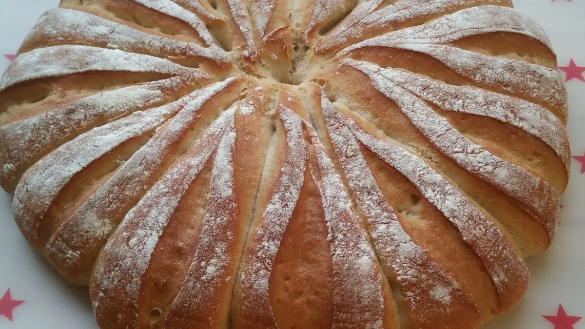 Ein runder Br&ouml;tchen-Kranz liegt auf einem Holzbrett, leicht mit Puderzucker best&auml;ubt, in einer hellen K&uuml;che.