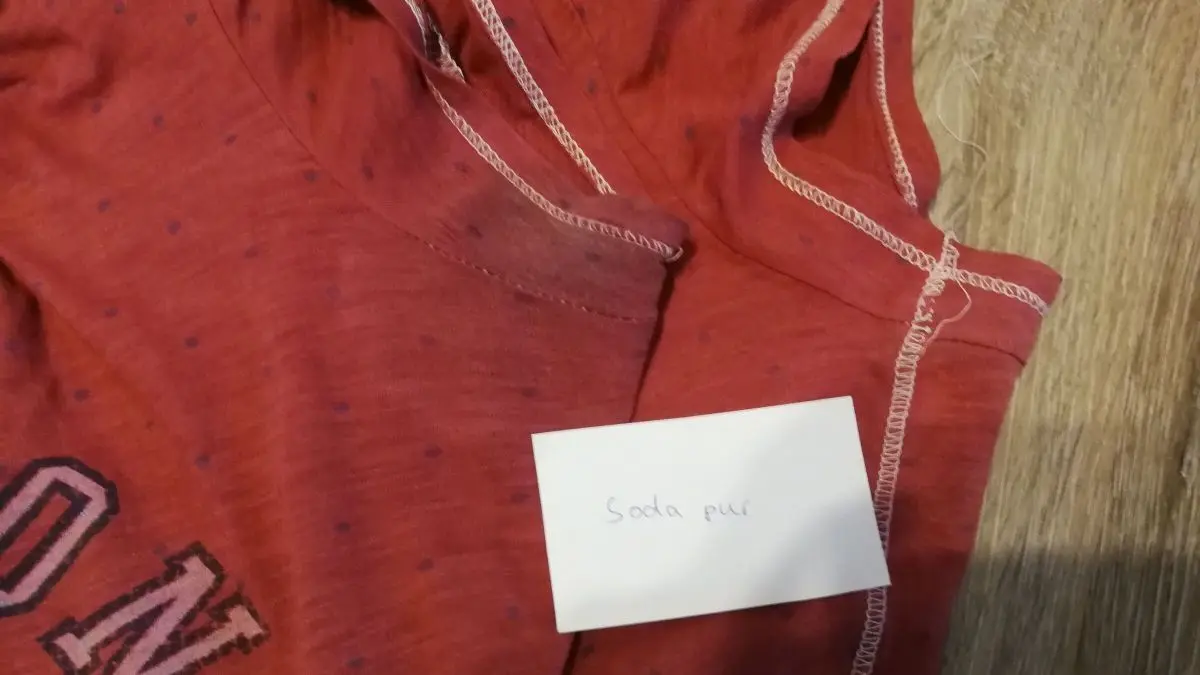 Ein rotes T-Shirt mit Deoflecken liegt auf einem hellen Untergrund, daneben befindet sich ein kleiner Notizzettel.