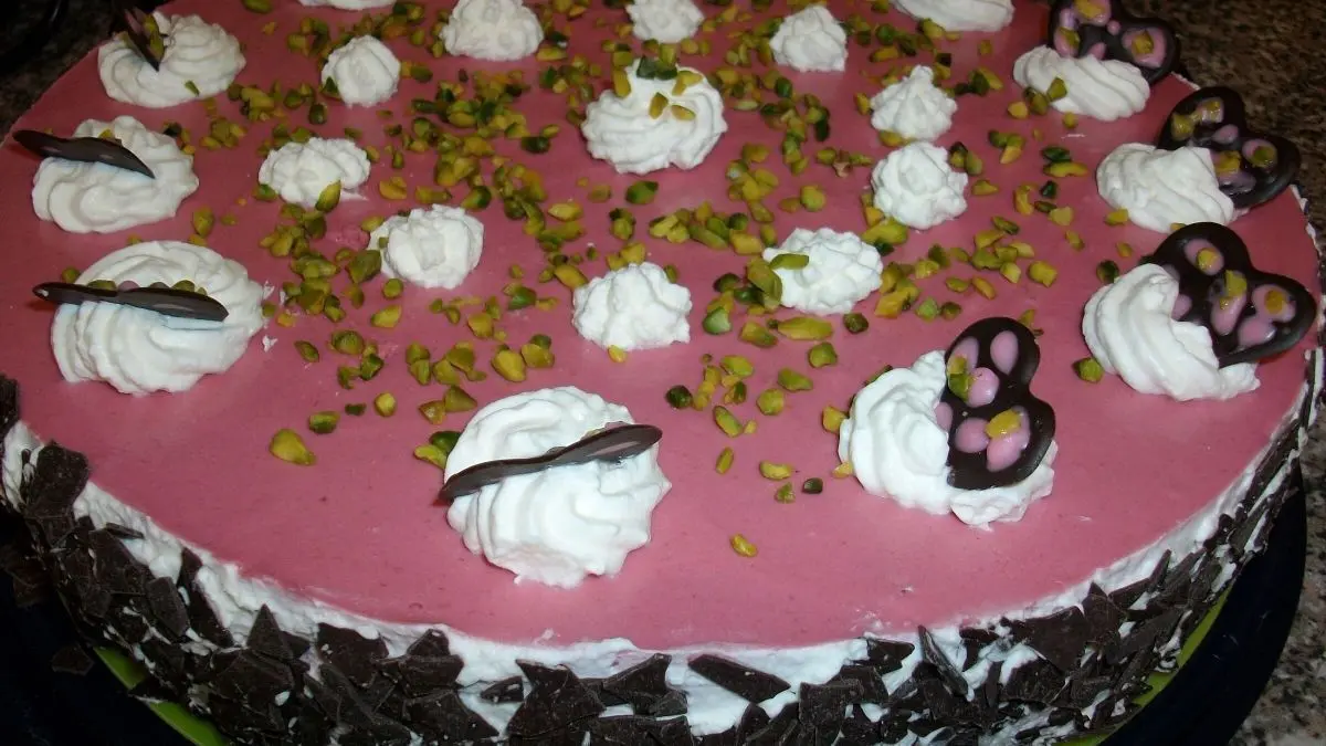 Eine Himbeertorte mit Mascarpone und Sahne liegt zentral auf einer gr&uuml;nen Platte, dekoriert mit Sahneh&auml;ubchen und Pistazien.