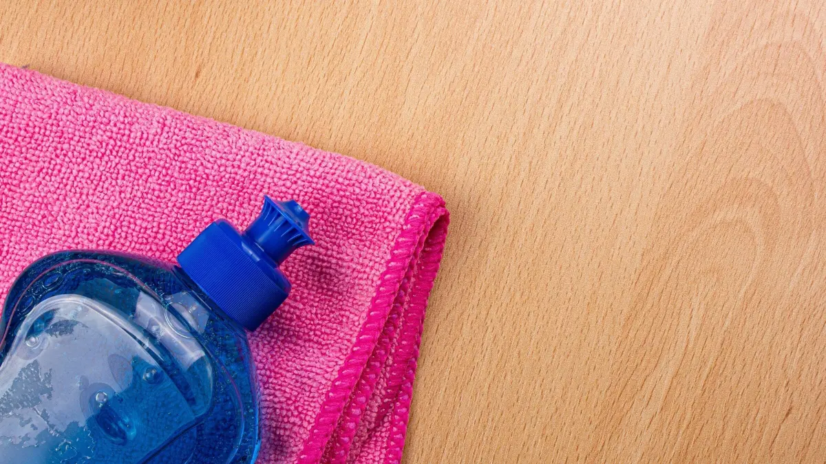 Ein blauer Sp&uuml;lmittelflasche liegt auf einem pinken Tuch auf einem hellen Holztisch, bereit zur Nutzung.