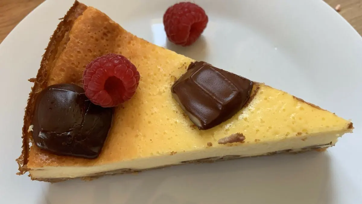 Ein St&uuml;ck K&auml;sekuchen mit Keksboden liegt auf einem wei&szlig;en Teller. Es ist mit Kinderriegeln und zwei Himbeeren garniert.