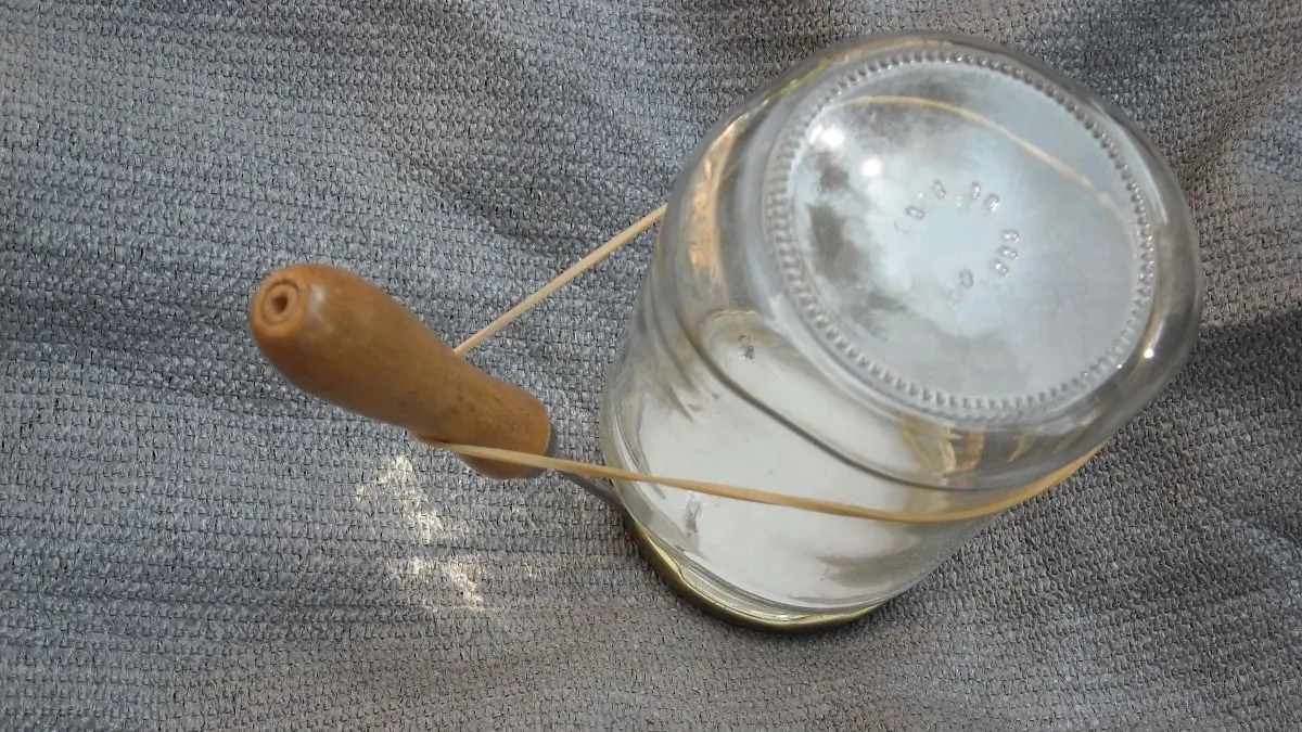 Ein Twist-Off-Glas liegt auf einem grau gemusterten Untergrund, w&auml;hrend ein Holzwerkzeug den Deckel m&uuml;helos &ouml;ffnet.