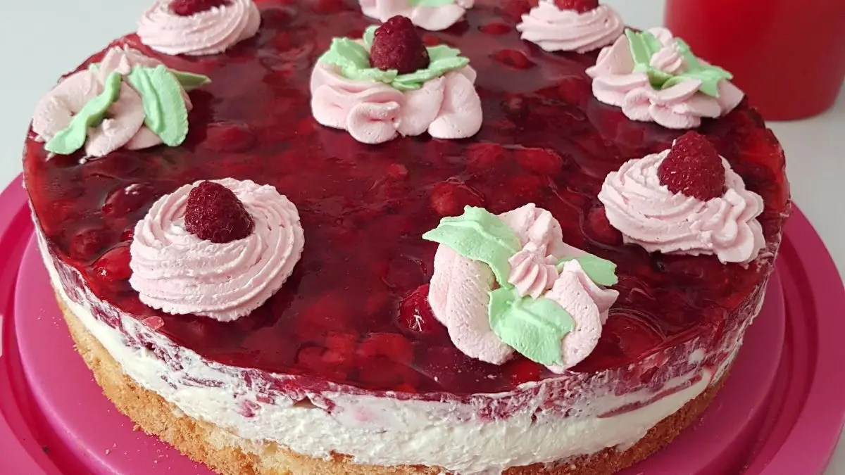 Eine saftige Himbeertorte mit einer Schmand-Sahne-F&uuml;llung steht auf einem rosa Teller, dekoriert mit Sahnetupfen und frischen Himbeeren.