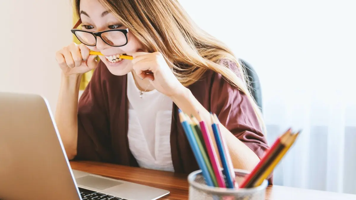 Eine konzentrierte junge Frau mit einer Brille bei der Arbeit am Laptop, umgeben von Buntstiften, schaut frustriert.