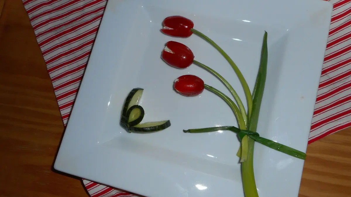 Rote Tomatentulpen stehen auf einem wei&szlig;en Teller, umgeben von einem gr&uuml;nen Schnittlauchst&auml;ngel, auf gestreiftem Untergrund.