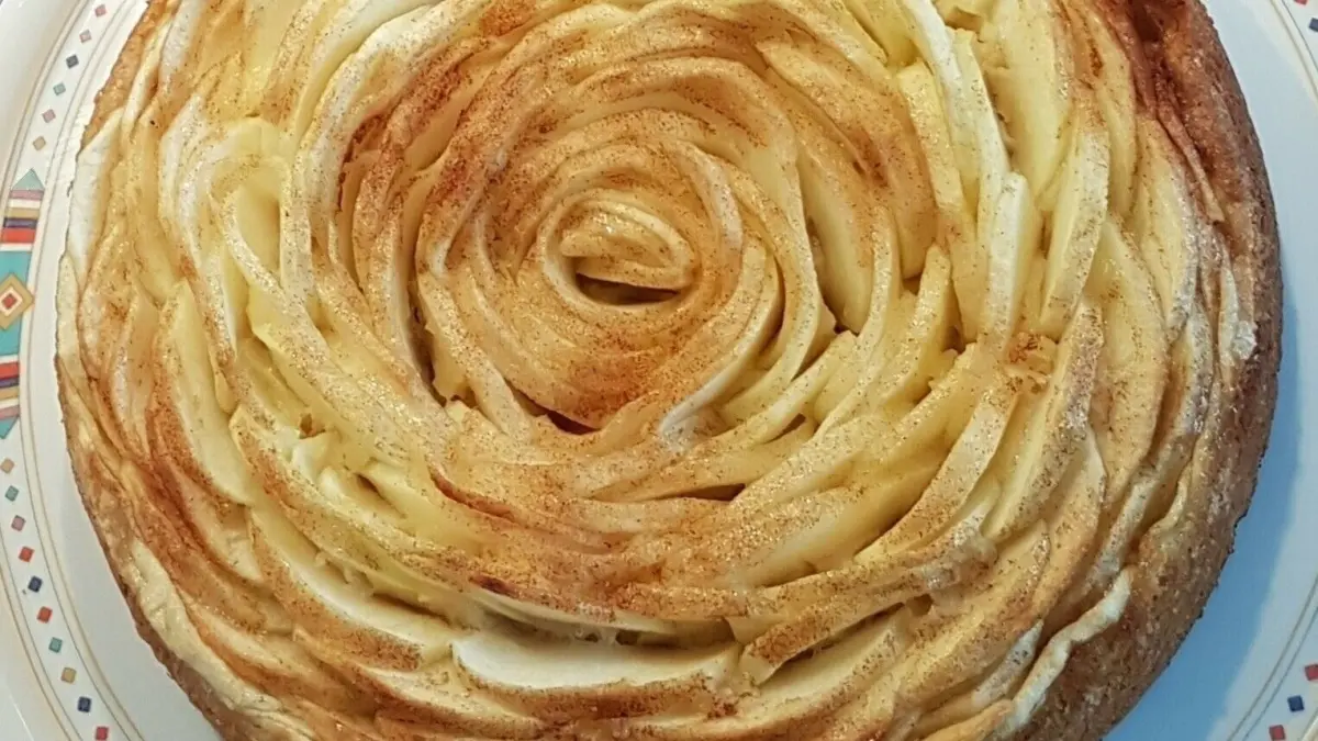 Eine kunstvoll dekorierte Apfelkuchentorte, geformt wie eine gro&szlig;e Rosenbl&uuml;te, ruht auf einem bunten Teller.