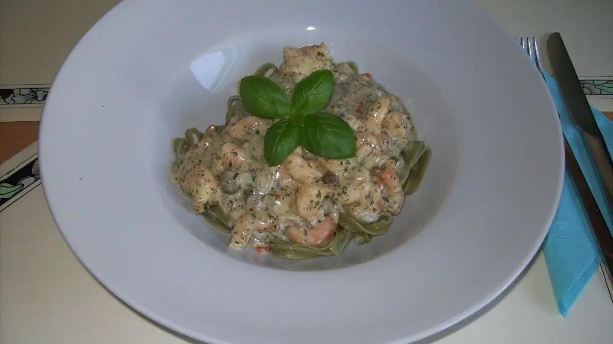 Ein Teller mit gr&uuml;nen Bandnudeln, bedeckt mit Scampi in Kr&auml;uter-Sahne, garniert mit frischem Basilikum, auf einem Tisch.