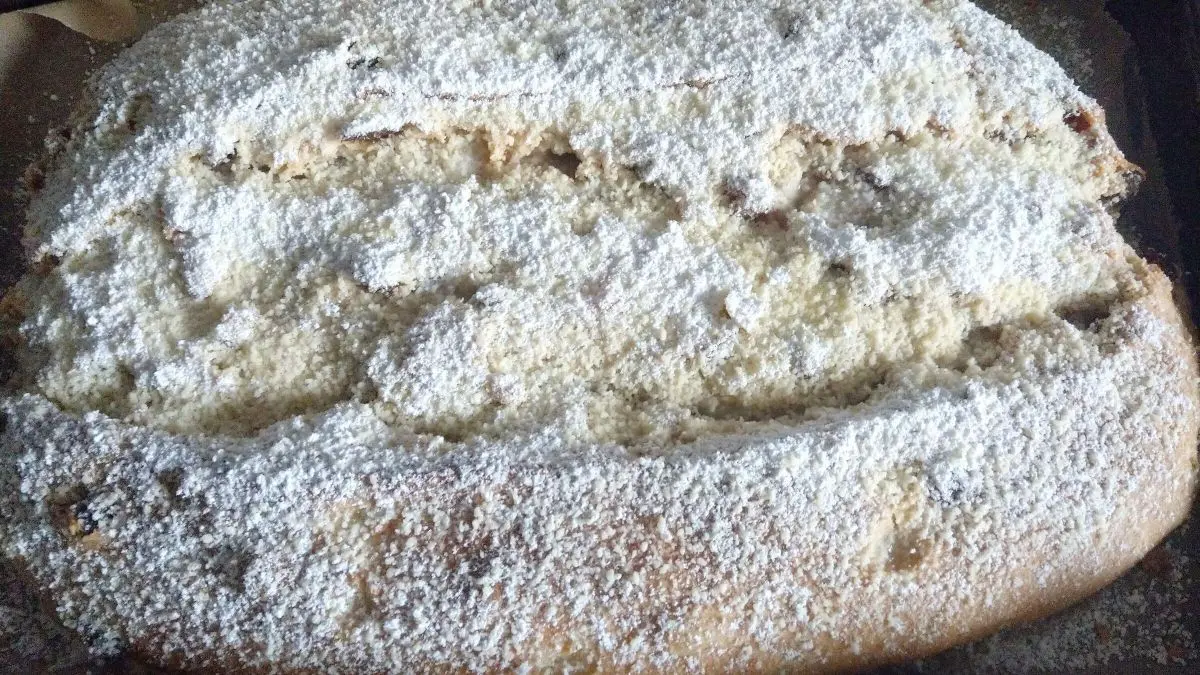 Ein frisch gebackener Butter-Rosinen-Stollen liegt auf einer Backplatte, best&auml;ubt mit Puderzucker und umgeben von Backpapier.
