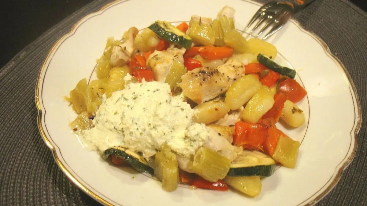 Ein Teller zeigt Ofengem&uuml;se mit Gnocchi und H&auml;hnchen-Geschnetzeltem, garniert mit einer cremigen Sauce auf einem grauen Platz.