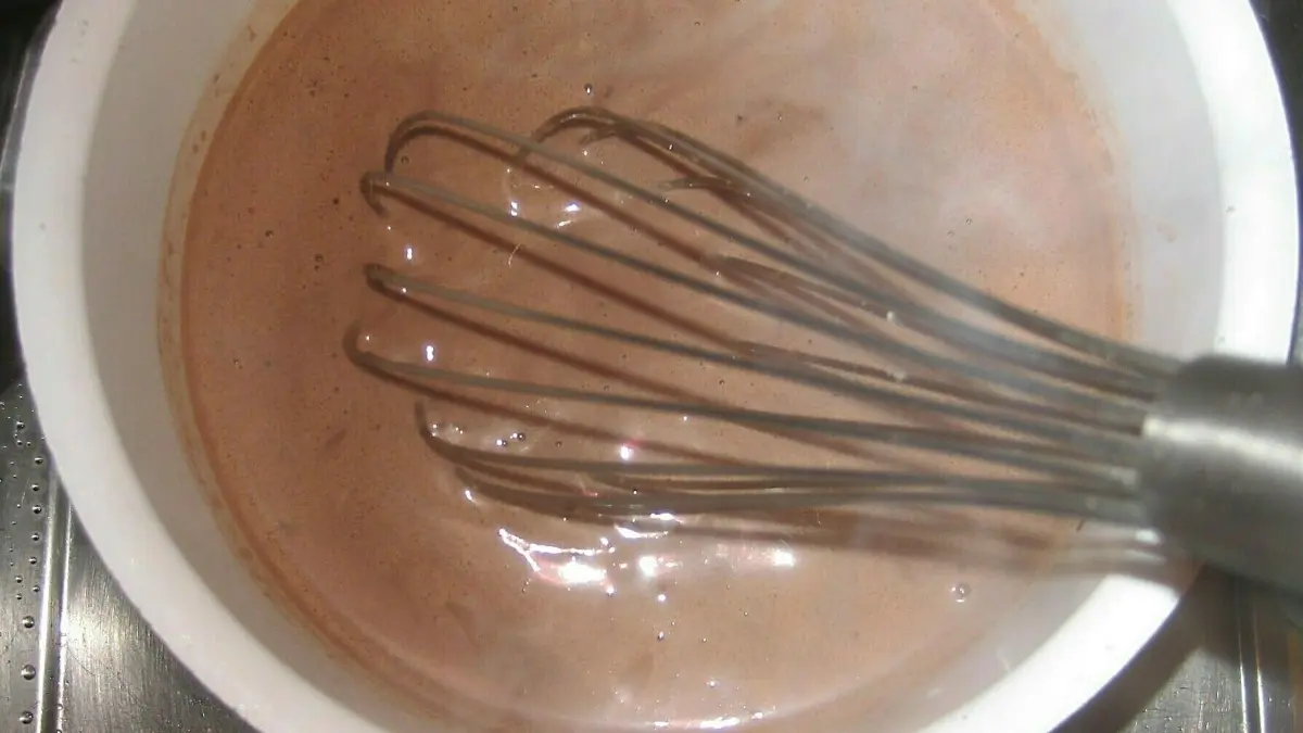 In einem wei&szlig;en Topf wird schokoladiger Pudding mit einem Schneebesen anger&uuml;hrt, w&auml;hrend Dampf aufsteigt.