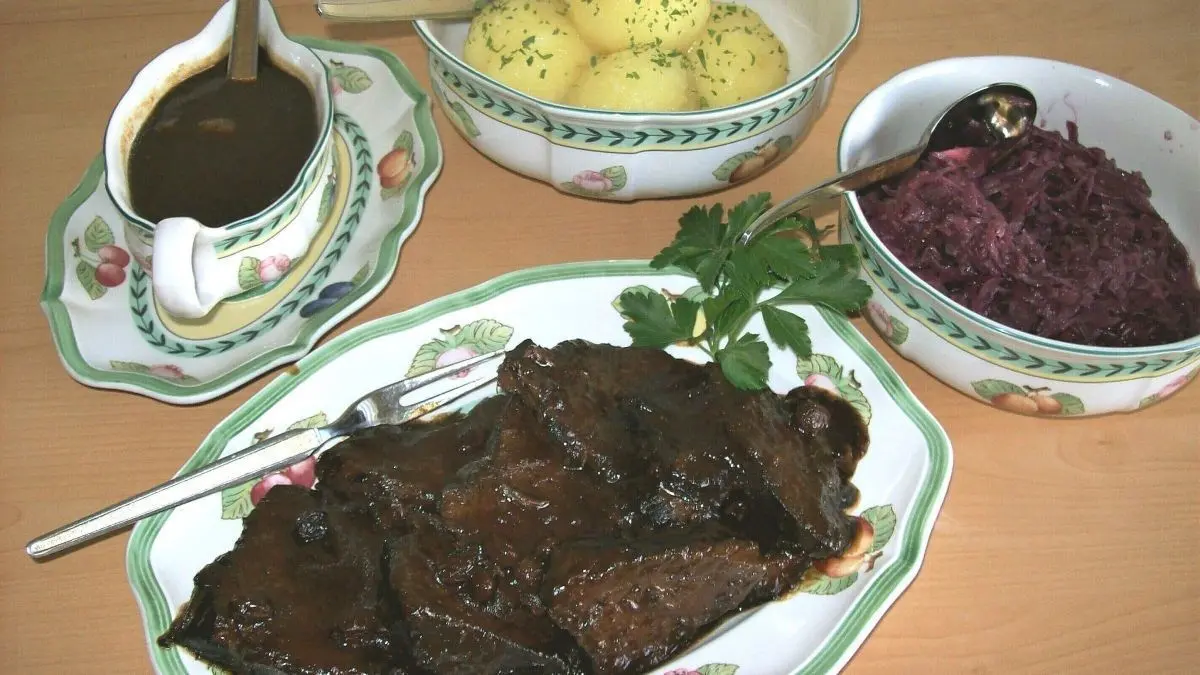 Ein saftiger Sauerbraten liegt auf einem Teller, umgeben von roten Kohl, Kartoffeln und So&szlig;e in eleganten Sch&uuml;sseln.