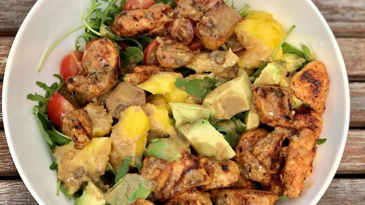 Eine bunte Salat Bowl mit H&auml;hnchenst&uuml;cken, Avocado und Erdnuss-Dressing, umgeben von frischen Tomaten und Holzoberfl&auml;che.