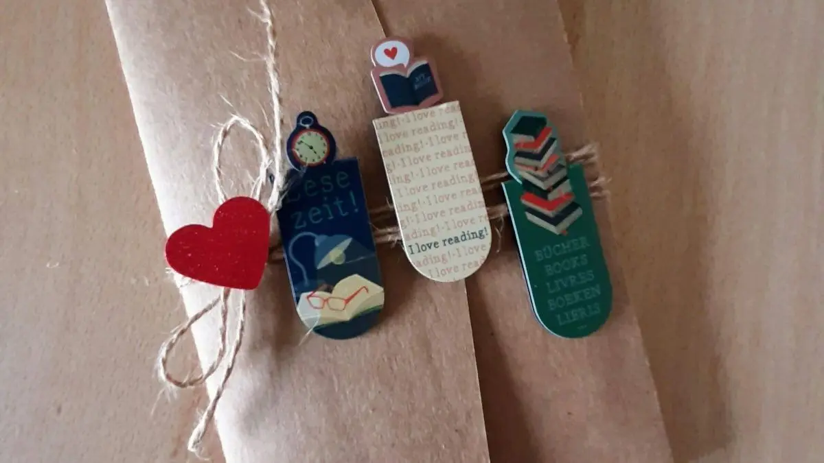 Eine dekorative Karte aus braunem Papier, liebevoll verziert mit Lesezeichen und einem roten Herz, liegt auf einem Tisch. Transkription: "Diese Zeit! I love reading! B&uuml;cher leben."