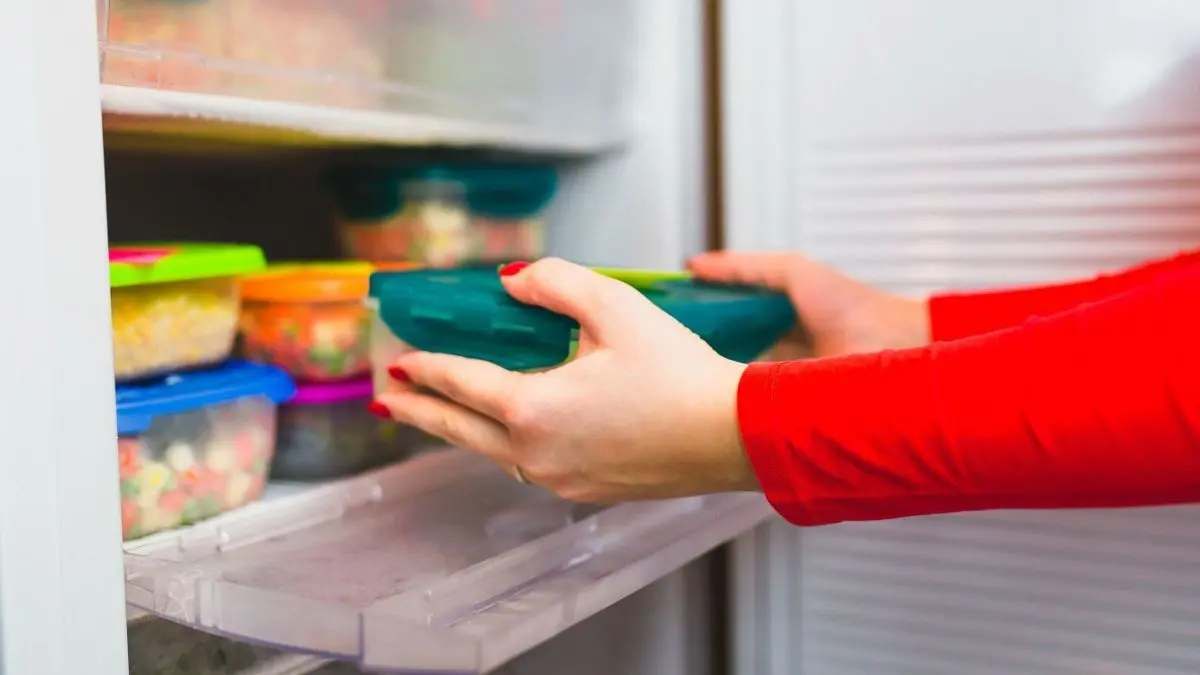 Eine Hand legt einen frischen Beh&auml;lter in einen gut organisierten Gefrierschrank mit bunten, beschrifteten Aufbewahrungsboxen.