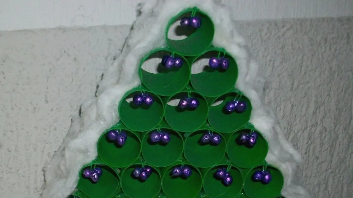 Ein gr&uuml;ner Weihnachtsbaum aus WC-Rollen mit lila Kugeln steht auf einer silbernen Oberfl&auml;che, umgeben von einer Wand.