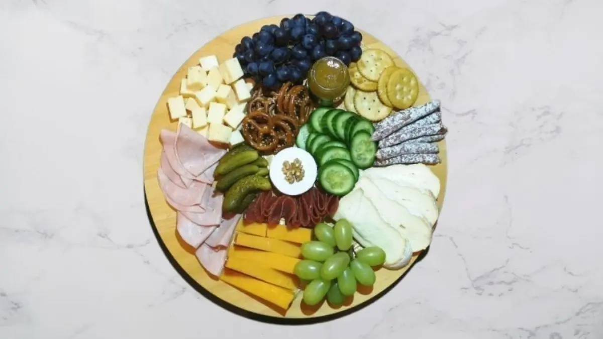 Eine runde Charcuterie-Board zeigt eine Vielzahl von Wurst, K&auml;se, Trauben, Gurken und Crackern, appetitlich angerichtet.
