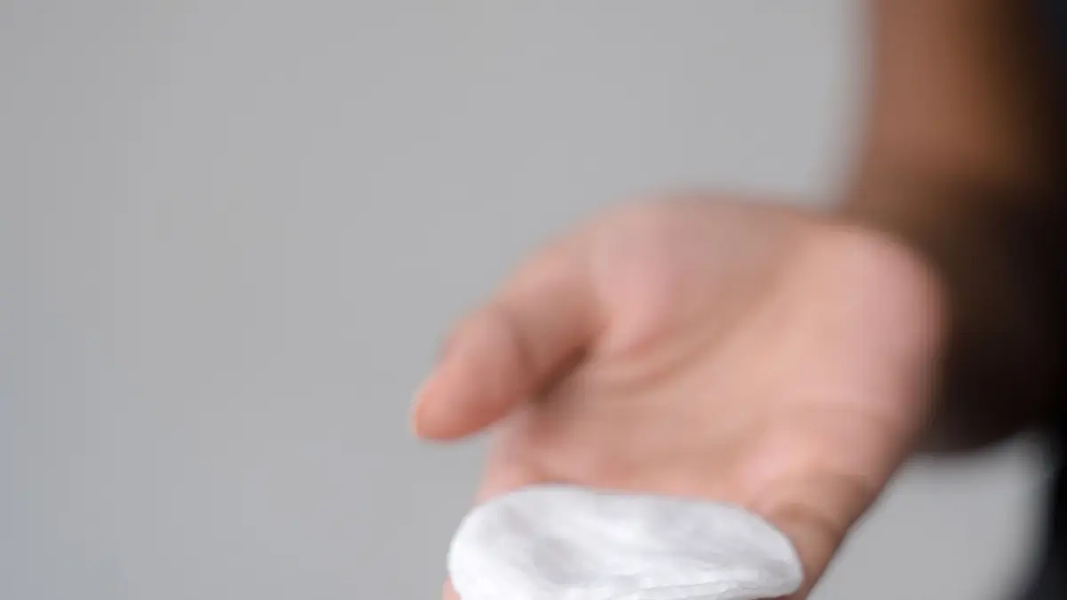 Eine Hand h&auml;lt ein saugf&auml;higes Wattepad, bereit zur Anwendung auf Pickeln, vor einem neutralen Hintergrund.