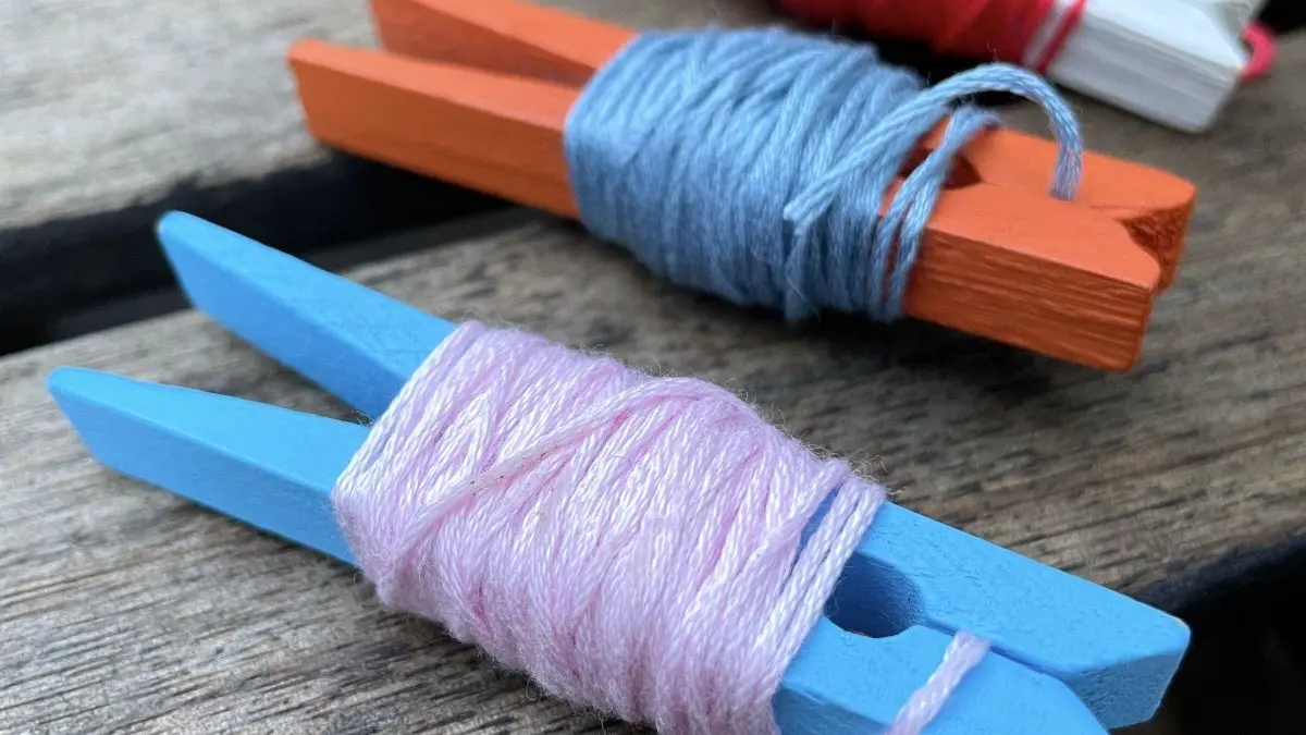 Bunte W&auml;scheklammern mit aufgereihtem Stickgarn liegen auf holzigem Untergrund und zeigen eine ordentliche Aufbewahrung.