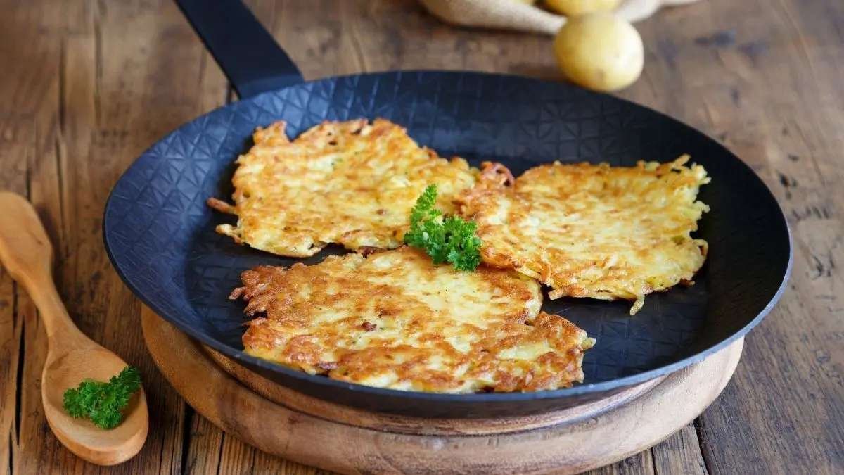 In einer schwarzen Pfanne liegen goldbraune Reibekuchen, umgeben von rohen Kartoffeln und einer Holzl&ouml;ffel.