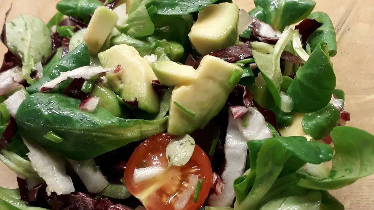 Feldsalat mit Avocado und Tomate wird frisch in einer Glasensch&uuml;ssel pr&auml;sentiert, auf einem Holzuntergrund.