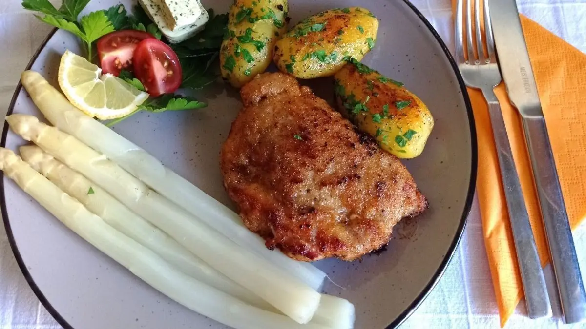 Ein knuspriges Cordon Bleu vom Schwein liegt auf einem Teller, umgeben von Spargel, Kartoffeln und frischem Gem&uuml;se.