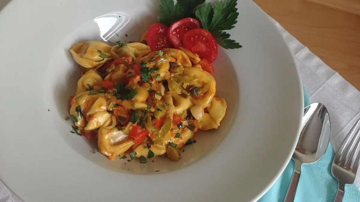 Tortelloni in scharfer So&szlig;e liegen ansprechend in einer wei&szlig;en Schale, umgeben von frischen Tomatenscheiben und Petersilie.