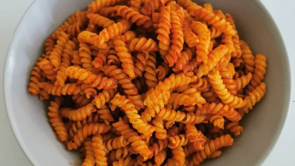 Eine Sch&uuml;ssel mit leuchtend orangefarbener Nudeln steht auf einem Tisch, dekoriert mit einer w&uuml;rzigen So&szlig;e.