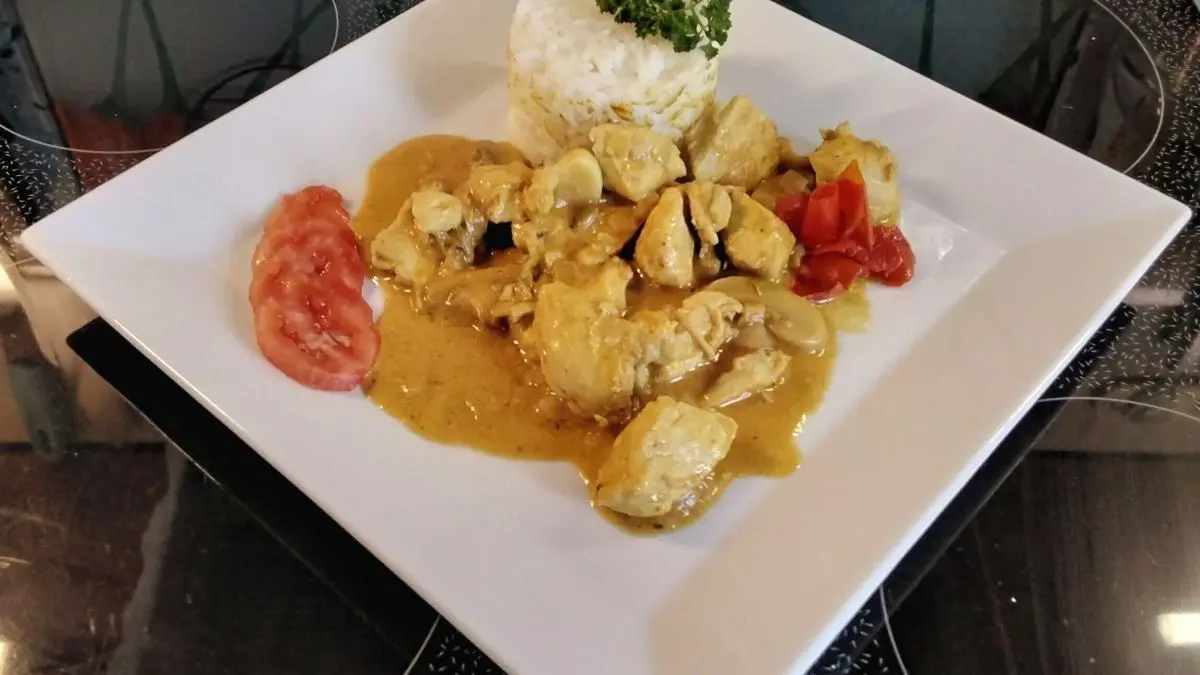 H&auml;hnchenbrust in Currysauce liegt neben Basmati-Reis und frischen Tomaten auf einem wei&szlig;en Teller in einer K&uuml;che.