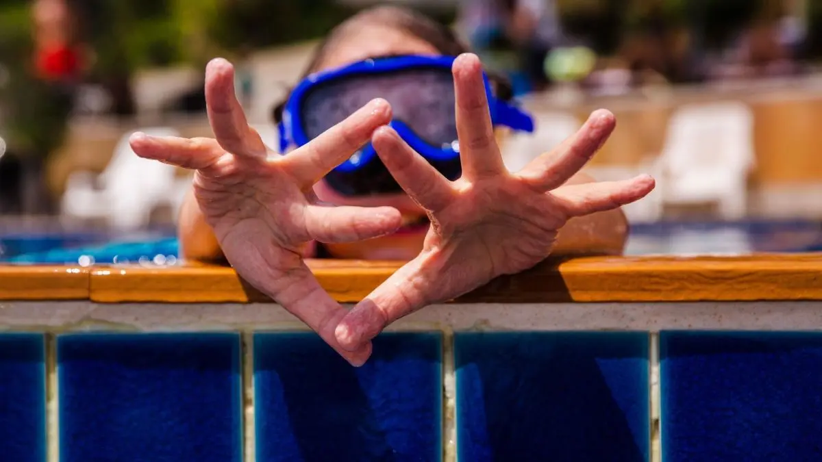 Fingers sind in Wasser sichtbar, w&auml;hrend eine Person mit Maske am Rand des Schwimmbeckens spielt und gestikuliert.