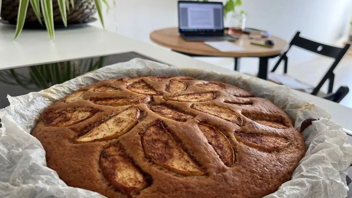 Ein saftiger Apfel-Joghurt-Kuchen mit Zimt ruht auf einem Tisch in einer modernen K&uuml;che, umgeben von Pflanzen und einem Laptop.