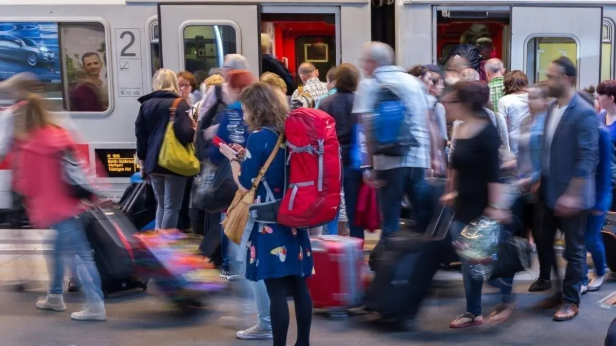 Eine Frau mit rotem Rucksack steht mit ihrem Handy in der Menge am Bahnhof, w&auml;hrend Reisende in den Zug einsteigen.