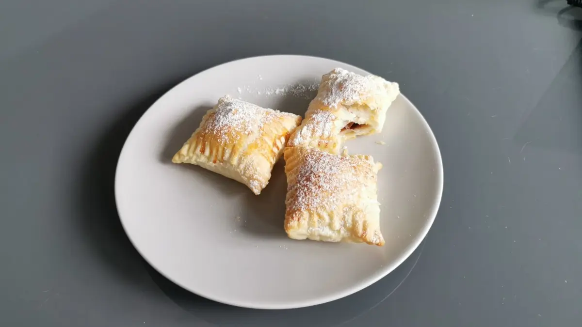 Drei Bl&auml;tterteigtaschen liegen auf einem wei&szlig;en Teller, bestreut mit Puderzucker, bereit zum Genie&szlig;en in einem hellen Raum.