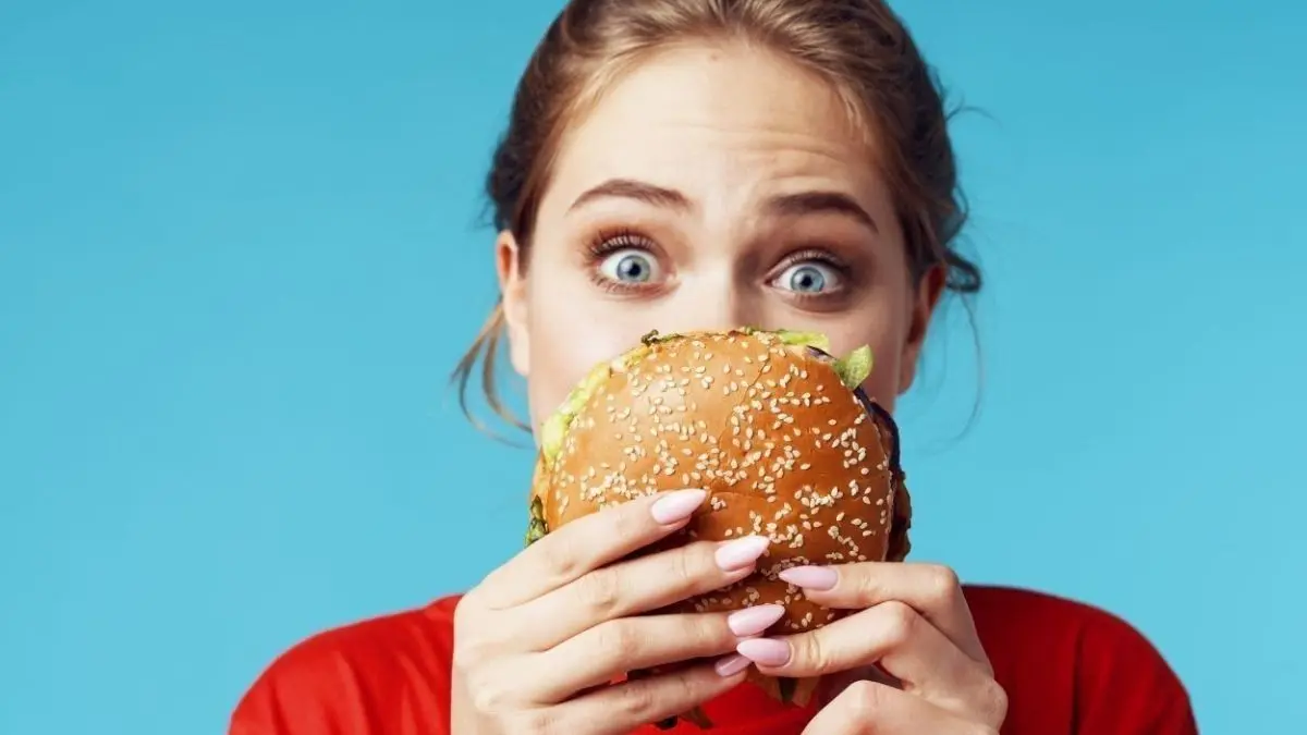 Eine junge Frau h&auml;lt einen Burger mit Sesambr&ouml;tchen vor ihrem Gesicht, w&auml;hrend sie aufgeregt in die Kamera schaut.