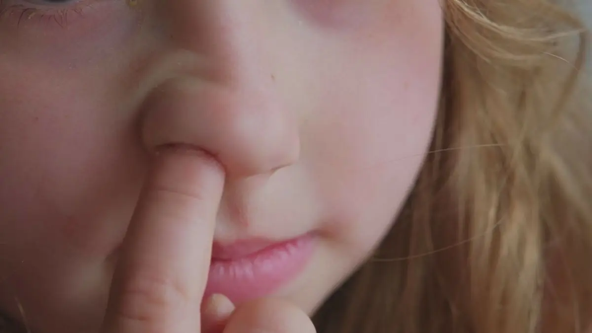 Ein Kind mit langen, blonden Haaren steckt den Finger in die Nase, w&auml;hrend es neugierig in die Kamera schaut.