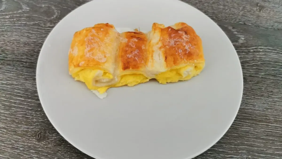 Auf einem wei&szlig;en Teller liegt ein gebackenes Puddingteilchen aus Bl&auml;tterteig, goldbraun und gef&uuml;llt mit cremiger Vanillef&uuml;llung.