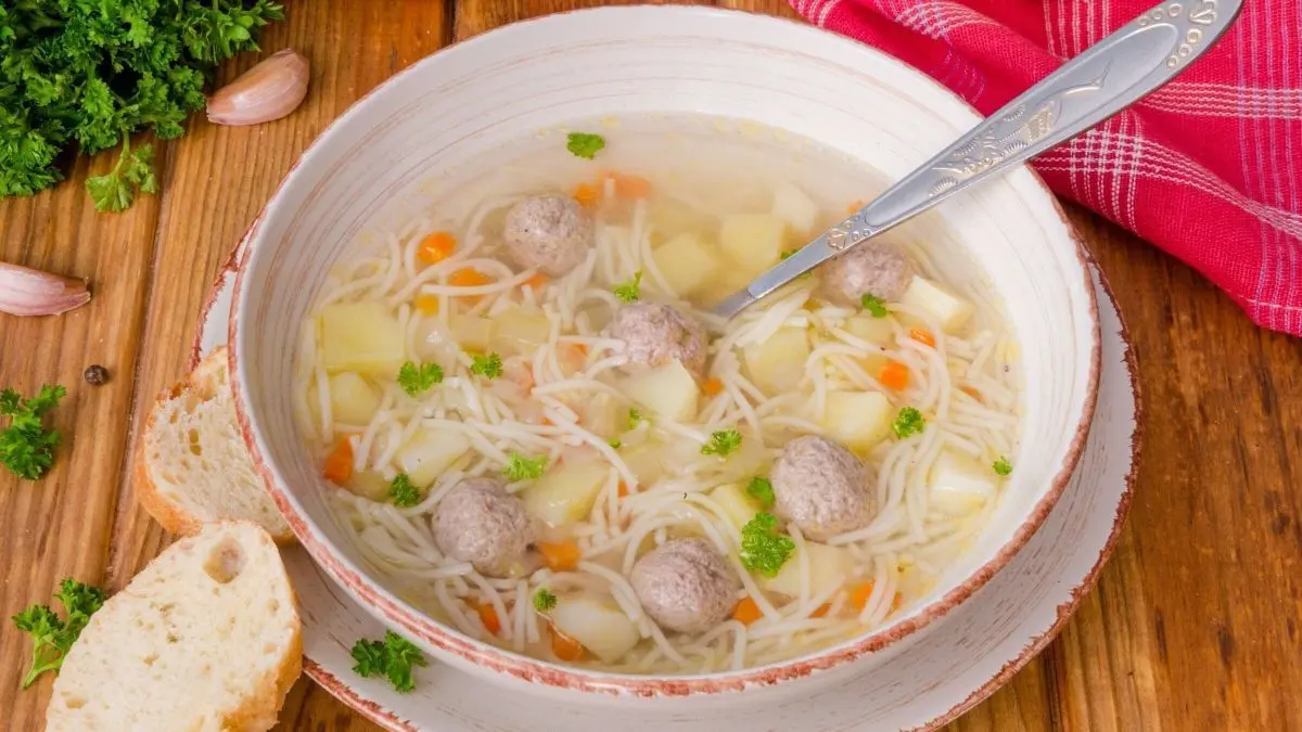 Eine Sch&uuml;ssel klare Suppe mit Einlagen wie Nudeln, Fleischb&auml;llchen und Gem&uuml;se steht auf einem Holztisch, umgeben von Brot und Kr&auml;utern.