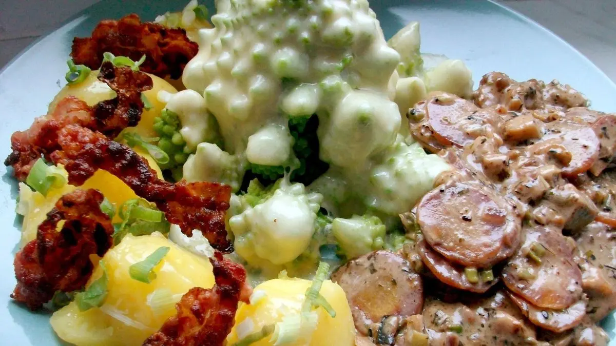 &Uuml;berbackener Romanesco liegt neben goldgelben Kartoffeln und knusprigem Speck auf einem blauen Teller.
