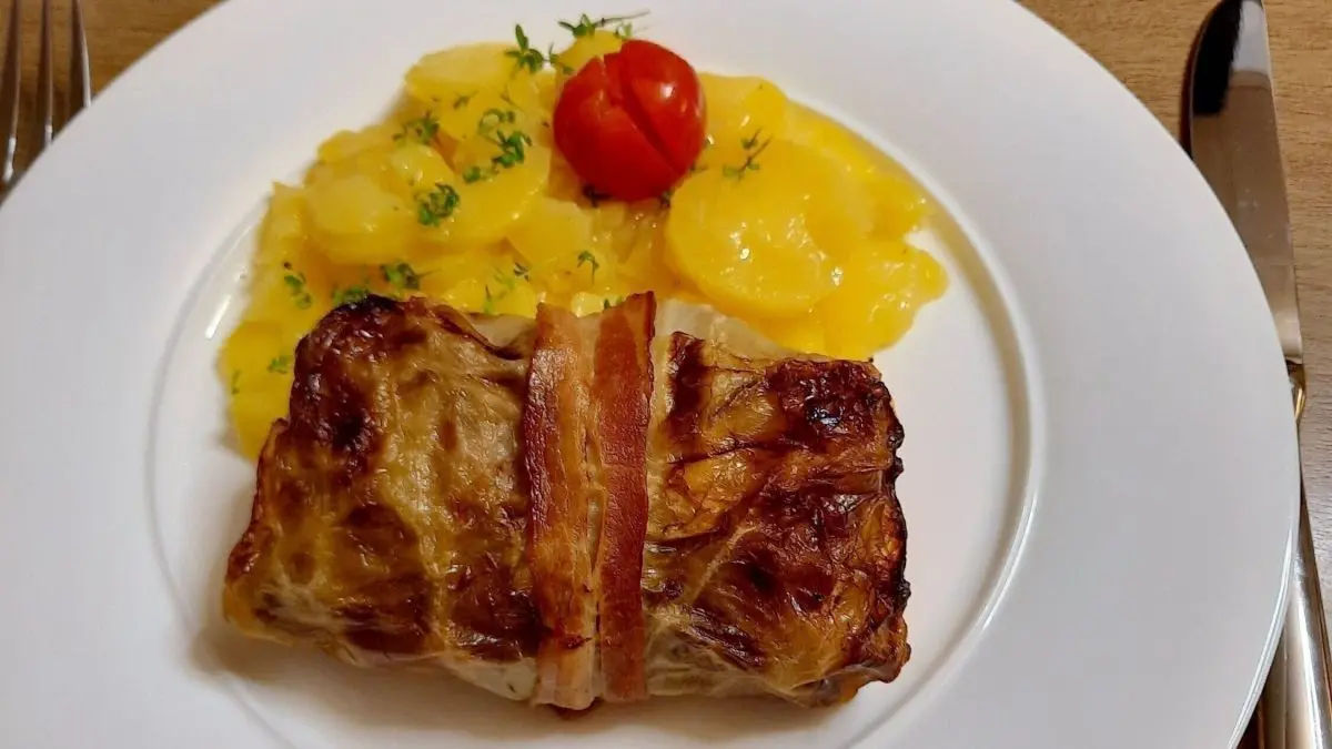 Ein saftiger Krautwickel mit Speck liegt auf einem wei&szlig;en Teller neben gelben Kartoffelscheiben und einer Kirschtomate.