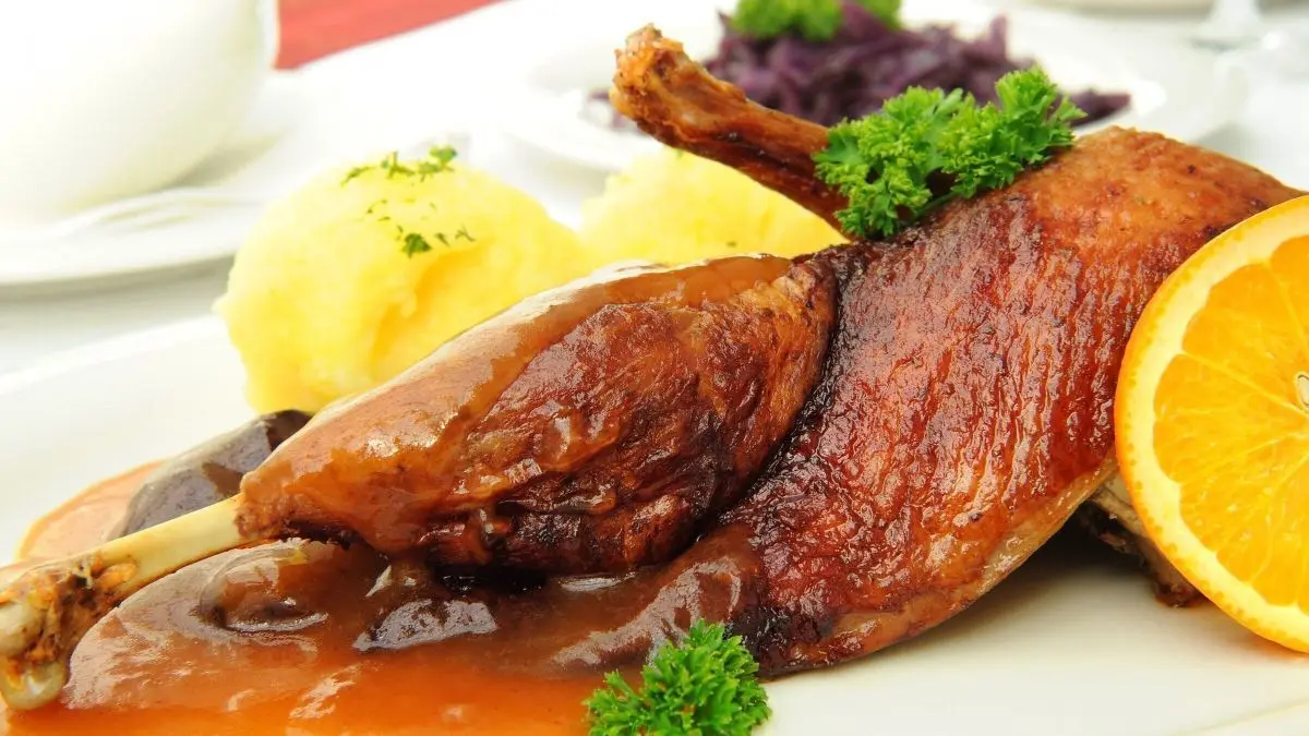 Entenbraten mit knuspriger Haut liegt auf dem Teller, umgeben von Serviettenkn&ouml;deln und einer Orangenscheibe.