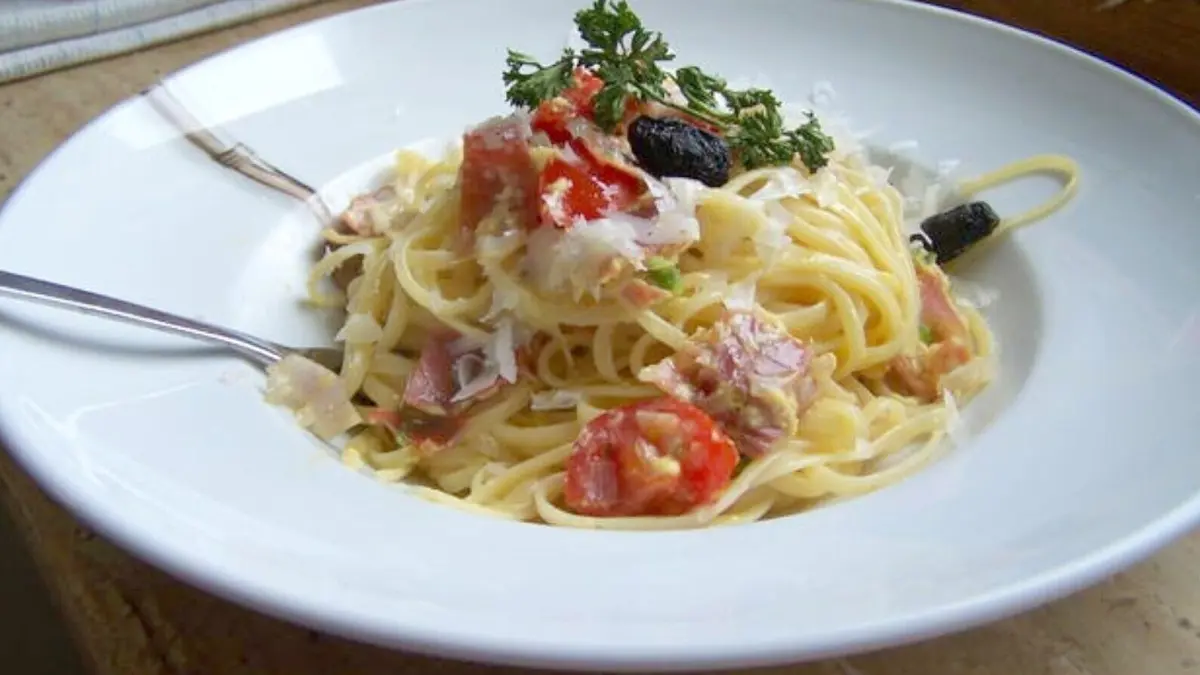 Ein Teller Spaghetti schwenkt mit Serrano-Schinken und Ei, verziert mit frischen Kr&auml;utern und Tomaten, vor einer hellen Kulisse.