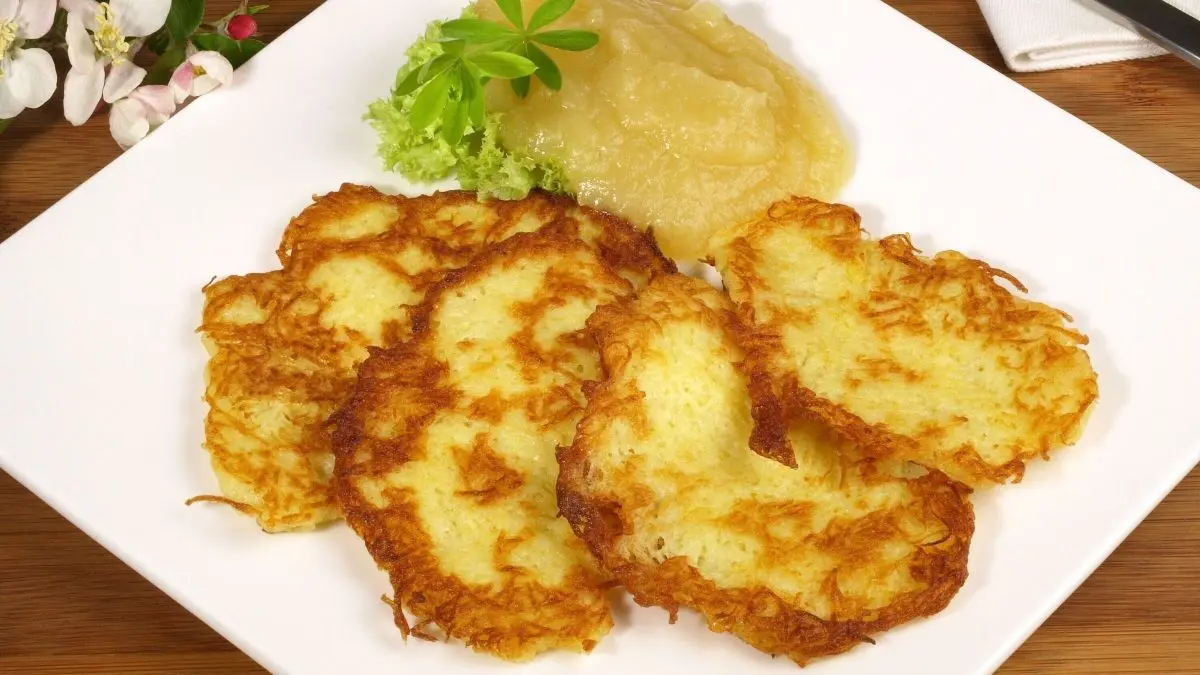 Knusprige Kartoffelpuffer liegen auf einem wei&szlig;en Teller, umgeben von Apfelmus und frischen Kr&auml;utern.