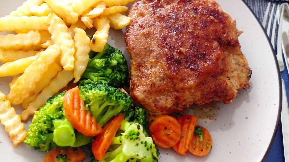 Ein gebratenes Cordon Bleu aus Schweinefleisch liegt auf einem Teller, umgeben von Pommes und buntem Gem&uuml;se.
