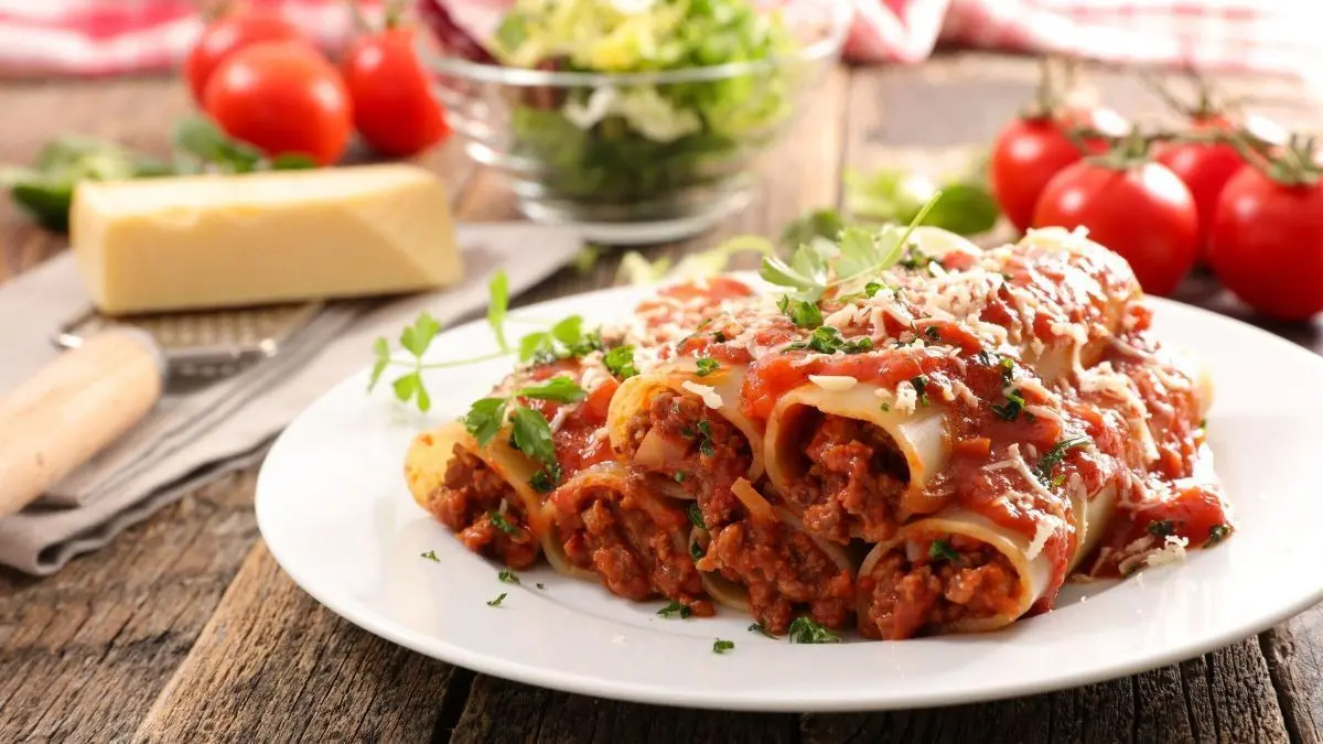 Cannelloni gef&uuml;llt mit Hackfleisch liegen auf einem wei&szlig;en Teller, umgeben von frischen Tomaten und Salat in einer rustikalen K&uuml;che.
