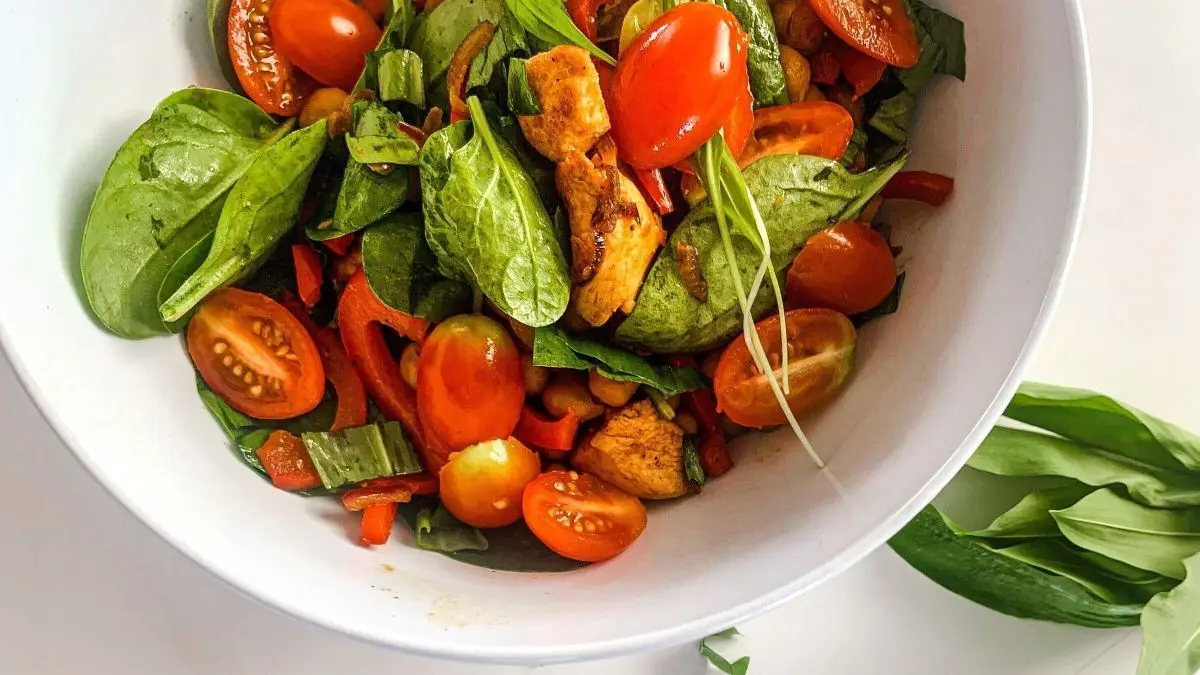 Ein bunter Kichererbsensalat mit H&auml;hnchen, Spinat und frischen Tomaten wird in einer Sch&uuml;ssel pr&auml;sentiert, umgeben von B&auml;rlauch.
