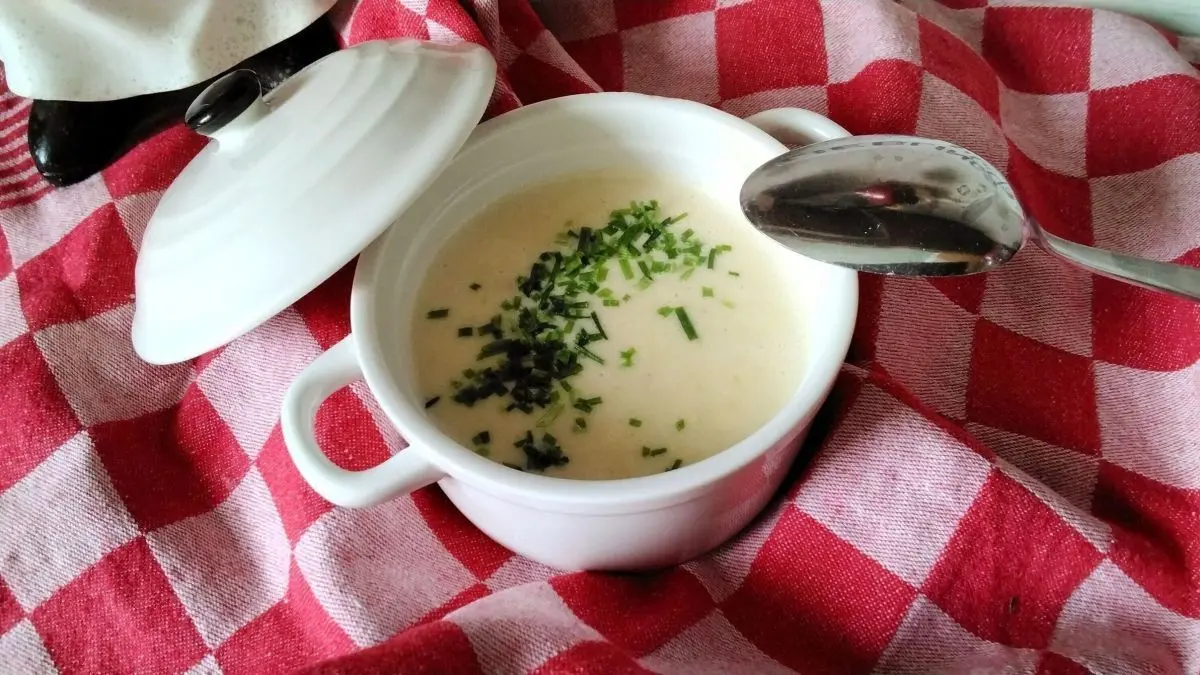 Eine wei&szlig;e Sch&uuml;ssel mit cremiger Spargelsuppe, garniert mit frischem Schnittlauch, steht auf einem rot-wei&szlig;en karierten Tuch.