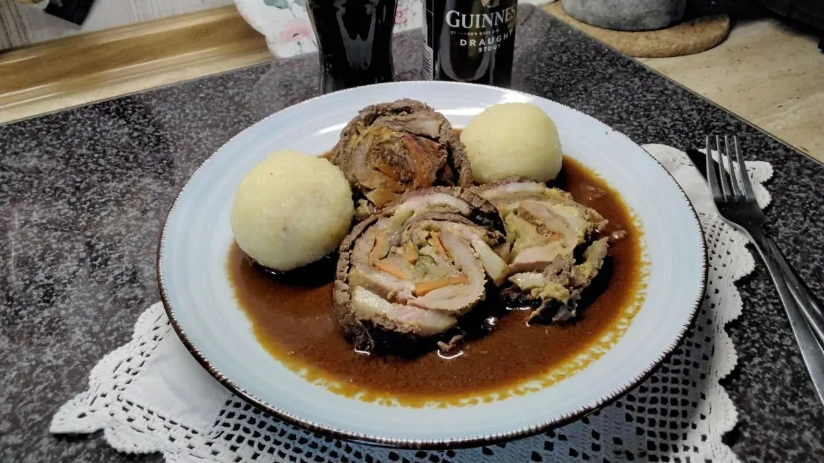 Eine gro&szlig;e Rinderroulade liegt auf einem Teller in Sauce, daneben zwei Kartoffelkn&ouml;del und ein Glas Guinness-Bier.